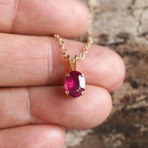 Könnte beinhalten: Eine goldfarbene Halskette mit einem kleinen, ovalförmigen Rubinanhänger. Der Rubin hat eine tiefrote Farbe und ist in eine goldene Lünette gefasst. Die Kette ist filigran und scheint aus Gold zu sein.