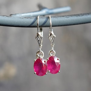 May include: A pair of silver drop earrings with oval, faceted ruby gemstones. The earrings have a decorative design at the top and a lever back closure. The rubies are a deep, vibrant red color.