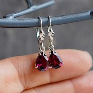 May include: A pair of silver drop earrings with pear-shaped, deep red gemstones. The earrings are suspended from decorative silver hooks. The gemstones are faceted, catching the light and displaying a rich, dark red color. The earrings are displayed against a neutral background.