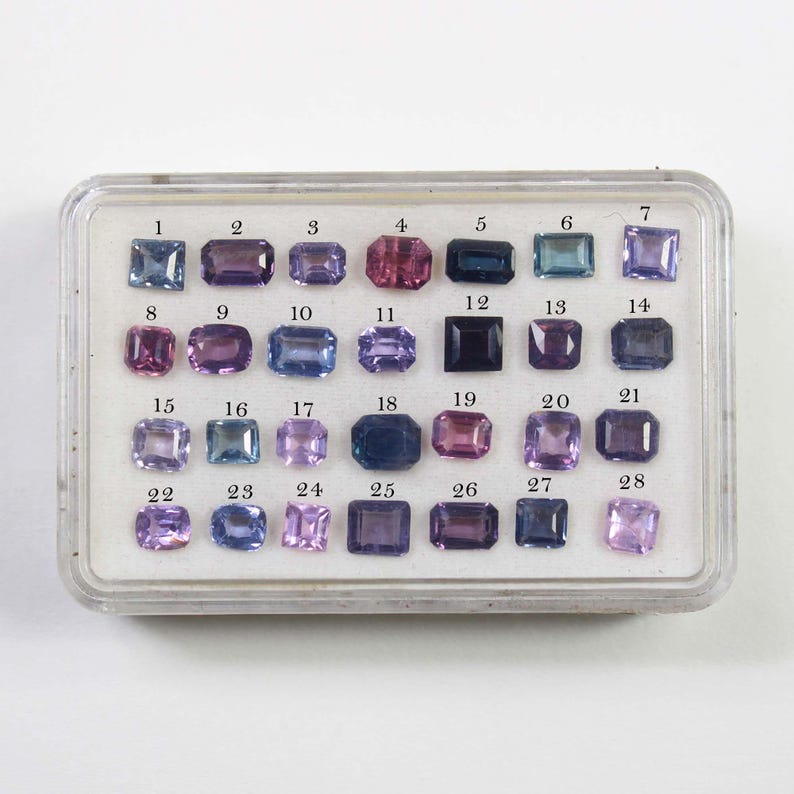 May include: A clear plastic case filled with 28 gemstones in various shapes and colors, including blue, purple, pink, and green. Each gem is numbered from 1 to 28, arranged in rows and columns.