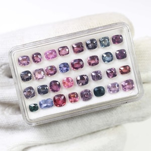 May include: A clear plastic case filled with a variety of colorful gemstones. The gemstones are square-cut and range in color from pink and purple to blue and green. The case is resting on a white surface.