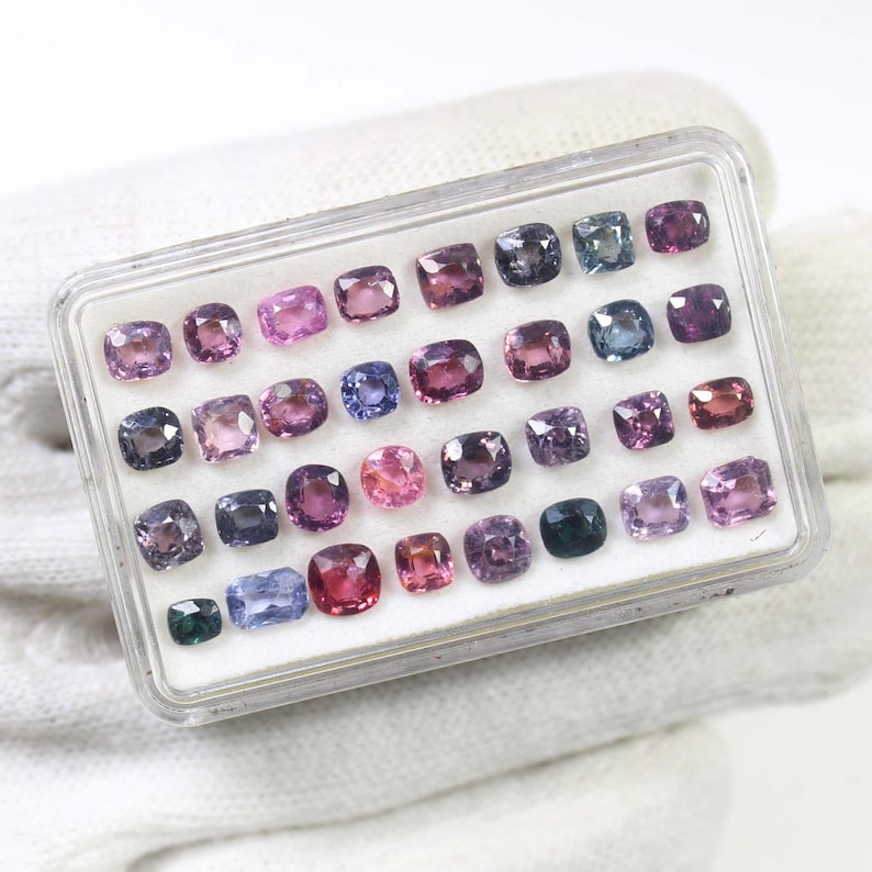 May include: A clear plastic case filled with a variety of colorful gemstones. The gemstones are square-cut and range in color from pink and purple to blue and green. The case is resting on a white surface.