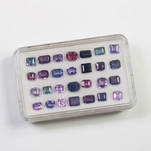 May include: A clear plastic case filled with an assortment of colorful gemstones. The gemstones are various shapes, including square, rectangular, and oval, and in shades of purple, blue, pink, and green. The case is on a white surface.