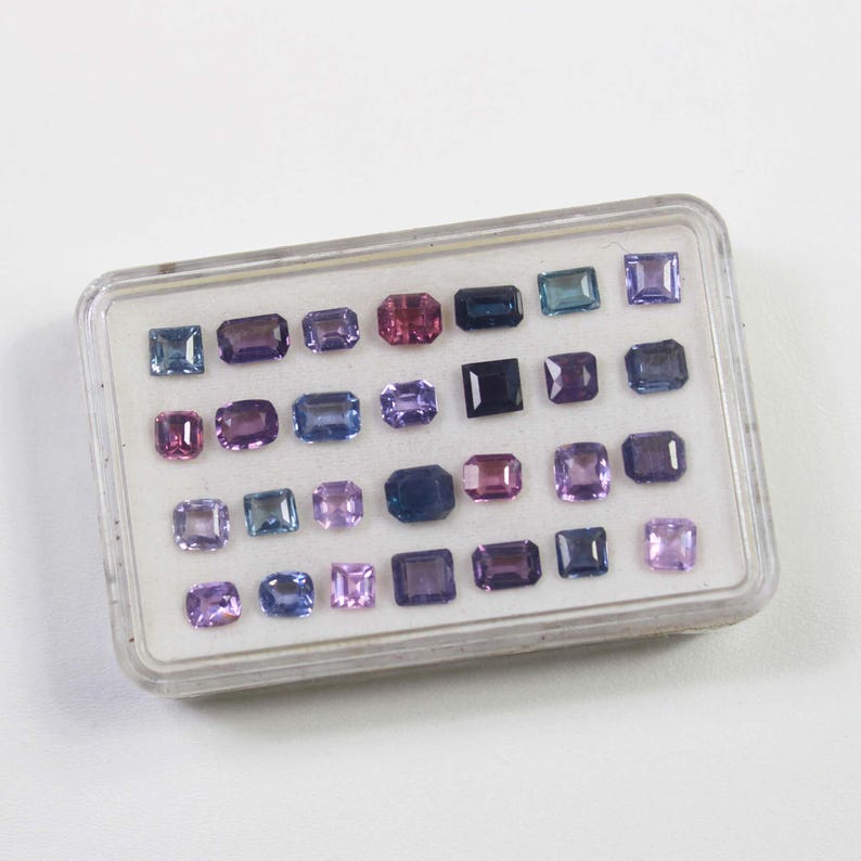 May include: A clear plastic case filled with an assortment of colorful gemstones. The gemstones are various shapes, including square, rectangular, and oval, and in shades of purple, blue, pink, and green. The case is on a white surface.