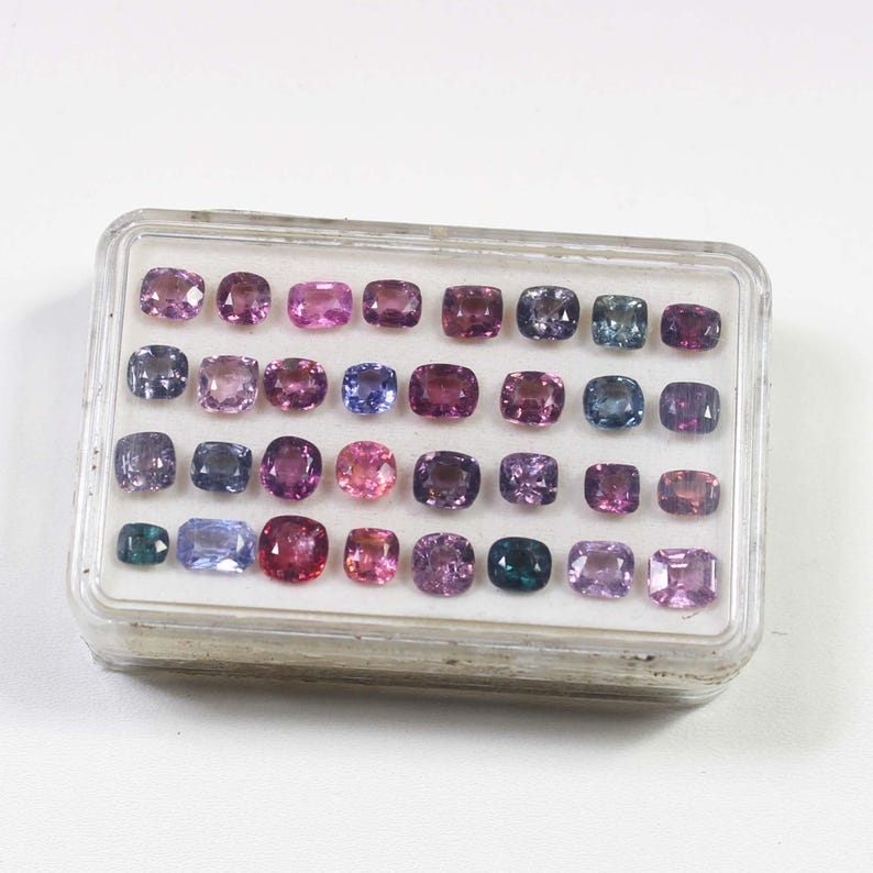 May include: A clear plastic case filled with an assortment of gemstones in various shades of purple, pink, blue, and green. The gemstones are cushion-cut and arranged in rows. The case is on a white surface.