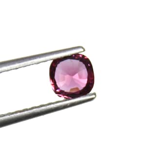 May include: A faceted, oval-shaped, pink gemstone held by silver tweezers. The gem has a polished surface and a vibrant, deep pink color. The tweezers are positioned against a bright white background.