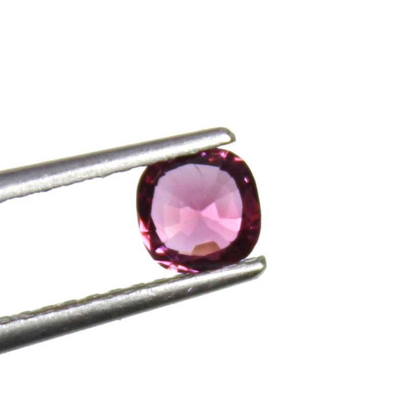 May include: A faceted, oval-shaped, pink gemstone held by silver tweezers. The gem has a polished surface and a vibrant, deep pink color. The tweezers are positioned against a bright white background.