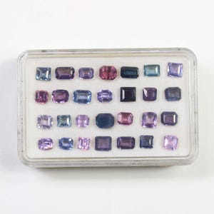 May include: A clear plastic case filled with an assortment of colorful gemstones. The gemstones are various shapes, including square, rectangular, and oval, and in shades of purple, pink, blue, and green. The case is on a white background.