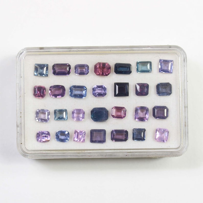 May include: A clear plastic case filled with an assortment of colorful gemstones. The gemstones are various shapes, including square, rectangular, and oval, and in shades of purple, pink, blue, and green. The case is on a white background.
