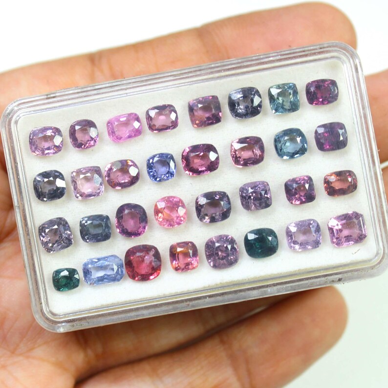 May include: A clear plastic case filled with a variety of colorful gemstones. The gemstones are square or rectangular, and come in shades of pink, purple, blue, and green. The case is held in a person's hand.