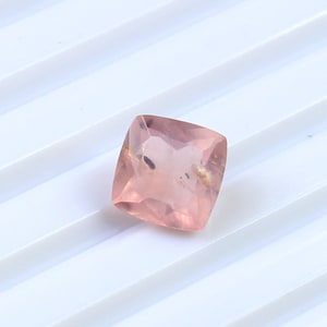 May include: A square-shaped, light pink gemstone with a slightly cloudy appearance. The gemstone has a smooth, polished surface.