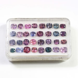May include: A clear plastic case filled with an assortment of gemstones in various shades of purple, pink, blue, and green. The gemstones are square or rectangular, and the case is on a white background.