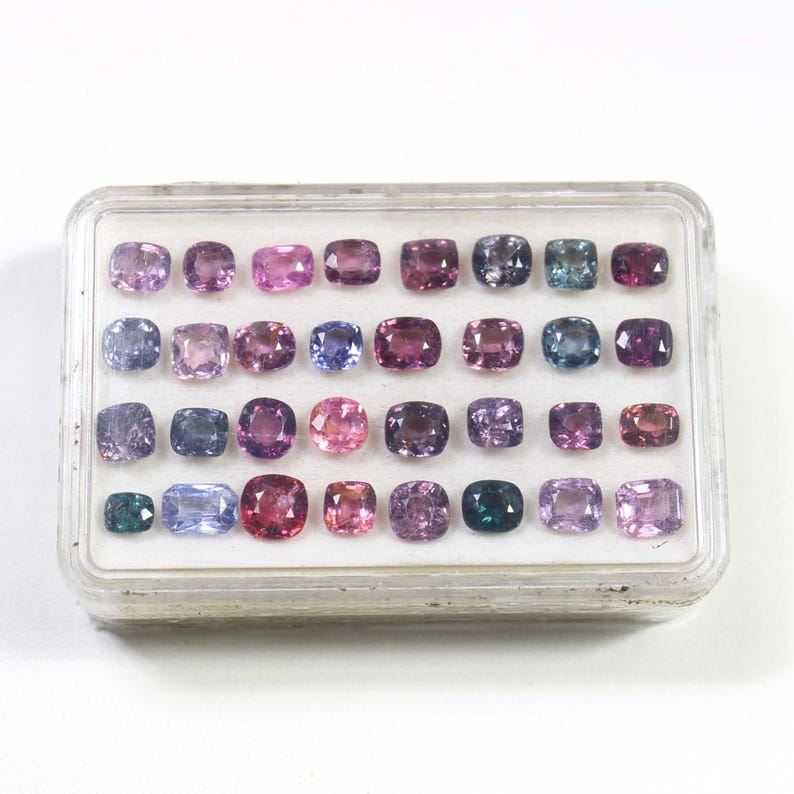 May include: A clear plastic case filled with an assortment of gemstones in various shades of purple, pink, blue, and green. The gemstones are square or rectangular, and the case is on a white background.