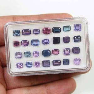 May include: A clear plastic case filled with an assortment of gemstones in various shapes and sizes. The gemstones are primarily shades of purple, pink, and blue. The case is held in a person's hand.