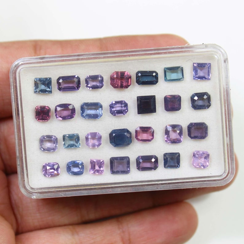 May include: A clear plastic case filled with an assortment of gemstones in various shapes and sizes. The gemstones are primarily shades of purple, pink, and blue. The case is held in a person's hand.