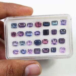 May include: A clear plastic case filled with an assortment of gemstones in various shapes and sizes. The gemstones are in shades of blue, purple, and pink. The case is held by a hand.