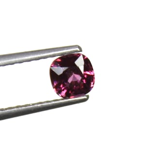 May include: A cushion-cut, purplish-red gemstone is held by a pair of tweezers against a white background. The gemstone has a faceted surface that reflects light, creating highlights and shadows. The tweezers are silver-colored.
