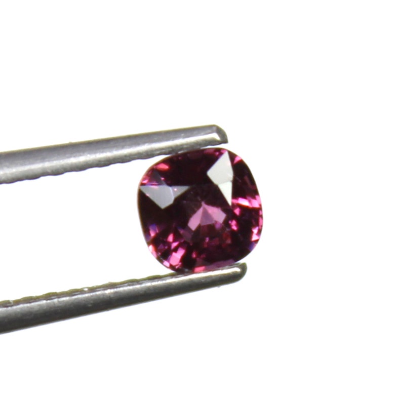 May include: A cushion-cut, purplish-red gemstone is held by a pair of tweezers against a white background. The gemstone has a faceted surface that reflects light, creating highlights and shadows. The tweezers are silver-colored.