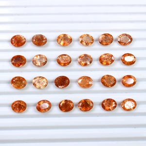 May include: Twenty-four oval-shaped sunstone gemstones in various shades of orange and brown. The stones are arranged in four rows of six on a white surface.