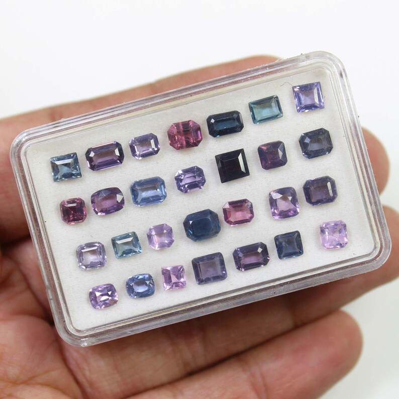 May include: A collection of various-sized gemstones in a clear plastic case. The gemstones are in shades of purple, blue, and pink. The gemstones are rectangular, square, and oval-shaped. The case is held in a person's hand.