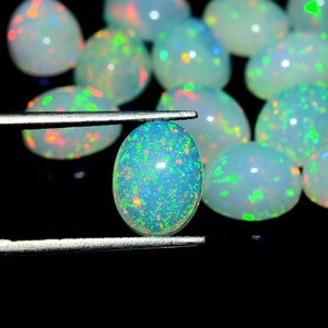 May include: A close-up of a white opal gemstone with green, blue, and orange flashes of color. The opal is oval-shaped and is being held by a pair of tweezers.