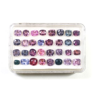 May include: A collection of gemstones in a clear plastic case. The gemstones are various shades of purple, pink, and blue. The stones are square or rectangular with rounded corners. The case is on a white background.