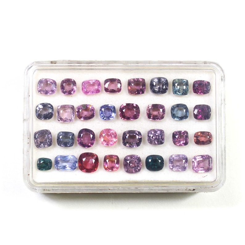 May include: A collection of gemstones in a clear plastic case. The gemstones are various shades of purple, pink, and blue. The stones are square or rectangular with rounded corners. The case is on a white background.