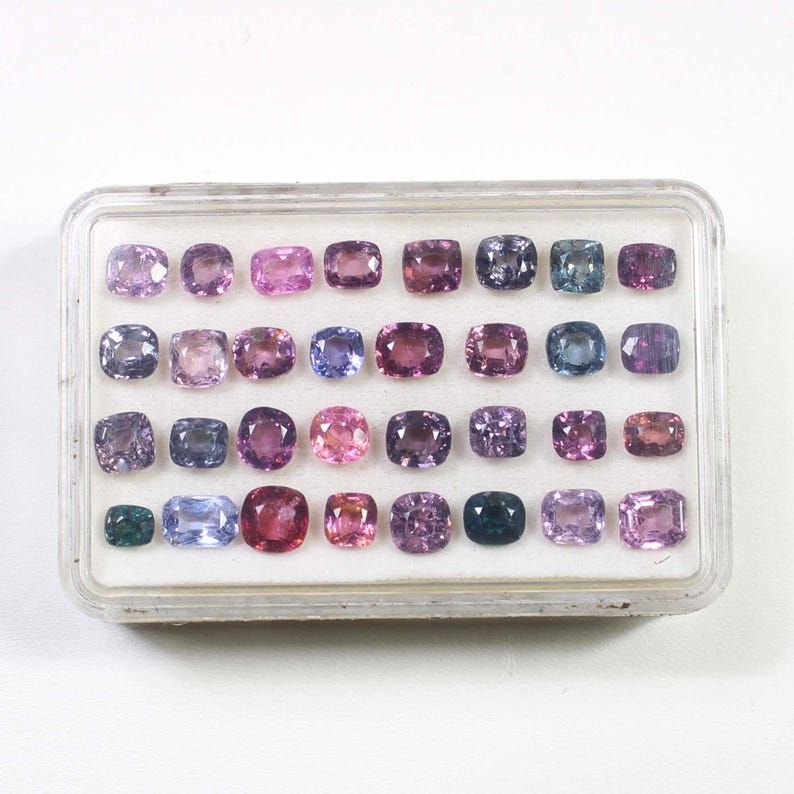 May include: A collection of cushion-cut gemstones in a clear plastic case. The gems display a variety of colors, including pink, purple, blue, and green. The arrangement is organized in rows and columns, showcasing the gems' facets.