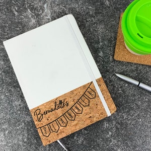 May include: A white notebook with a cork bottom. The cork has a black banner with the text "Bernadette's Planning Club" printed on it.