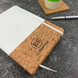 May include: A white and cork notebook with a black elastic band and the initials "ES" in a circle on the cork cover. The text "NOTEBOOK" is printed below the circle.
