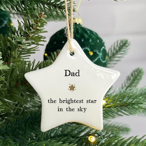 Op de afbeelding: Een witte, stervormige kerstversiering met het woord "Dad" en de zin "the brightest star in the sky" in het zwart gedrukt. De versiering hangt aan een touwtje van natuurlijk touw en wordt op een kerstboom getoond.