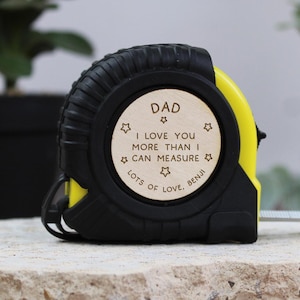 May include: A black and yellow tape measure with a wooden circular insert. The insert reads "DAD I LOVE YOU MORE THAN I CAN MEASURE LOTS OF LOVE, BENJI" with star accents. The tape measure is resting on a stone surface.