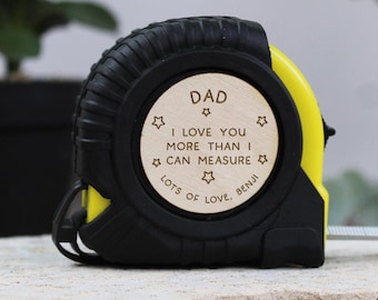 Puede incluir: Un flexómetro negro y amarillo con un inserto circular de madera. El inserto dice "DAD I LOVE YOU MORE THAN I CAN MEASURE LOTS OF LOVE, BENJI" con acentos de estrella. El flexómetro está sobre una superficie de piedra.