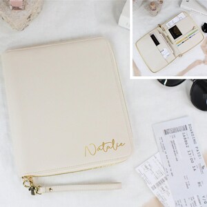May include: Cream-colored personalized travel wallet with a gold zipper and wrist strap. The wallet is embossed with the name "Natalie" in gold lettering. A gold pen is next to the wallet. Additional wallets in black, gray, pink, and brown are shown.