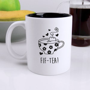 Peut inclure: Une tasse à café en céramique blanche avec un intérieur noir et une illustration en noir et blanc d'une tasse de thé avec un sachet de thé et le texte "FIF-TEA!"
