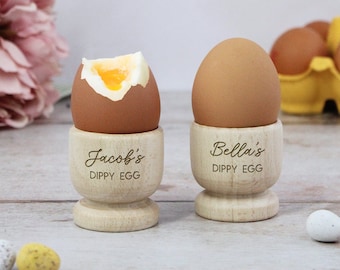 May include: Two wooden egg cups with personalized names "Jacob's Dippy Egg" and "Bella's Dippy Egg". One egg cup holds a boiled egg with the yolk showing.