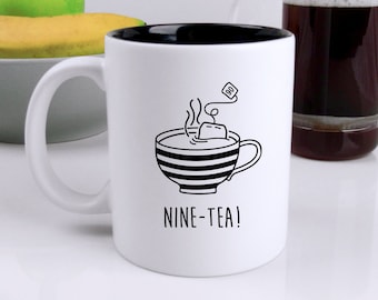 Peut inclure: Mug en céramique blanche avec intérieur noir, orné d'une illustration noire et blanche d'une tasse de thé avec un sachet de thé et le texte "NINE-TEA!". Une pomme verte et une banane sont dans un bol gris. Une cafetière à piston en verre est en arrière-plan.