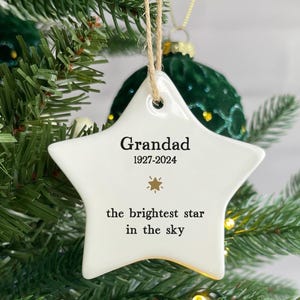 May include: A white star-shaped Christmas ornament with the text "Grandad 1927-2024" and "the brightest star in the sky." The ornament is suspended by a natural twine string and is displayed on a Christmas tree.