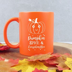 Peut inclure: Une tasse en céramique orange avec un dessin de citrouille blanc et le texte "Pumpkin Spice & Everything Nice". La tasse est sur une surface en bois avec des feuilles d'automne colorées.