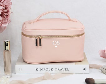 Op de afbeelding: Een lichtroze reistas voor make-up met een gouden rits en een handvat. De tas heeft de initialen "OS" en een klein hartje geprint op de voorkant. De tas ligt op een boek met de tekst "Kinfolk Travel" gedrukt op de kaft.