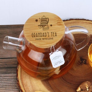 May include: A glass teapot with a wooden lid that says "Grandad's Tea Made With Love". The teapot is filled with tea and there is a tea bag hanging from the side. There are two chocolate chip cookies and a wooden spoon on a wooden serving tray.