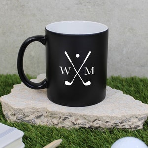 May include: Black coffee mug with a white interior, featuring a white golf-themed design. The design includes crossed golf clubs, a golf ball, and the initials "W M". The mug is on a stone surface with a golf ball and a notepad nearby.