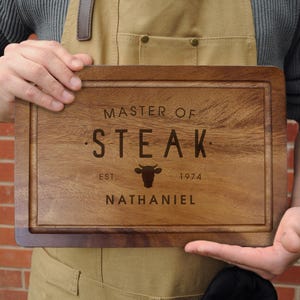 Puede incluir: Tabla de cortar rectangular de madera con acabado marrón oscuro. La tabla presenta el texto grabado "MASTER OF STEAK" con un gráfico de toro y el nombre "NATHANIEL". La tabla tiene una ranura para jugos alrededor del borde.