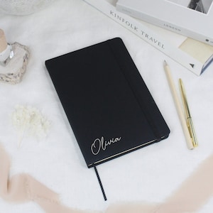 May include: A black leather-bound journal with a gold ribbon bookmark and the name "Olivia" embossed in silver on the front cover.