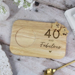 May include: A light wooden serving tray with a circular recess and the engraved text "40 AND Fabulous". Two star-shaped biscuits sit on the tray. A gold-coloured spoon rests nearby.