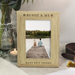 May include: A light oak picture frame with the text "BUDDY & ME" and "MANS BEST FRIEND" engraved. The frame displays a photo of a person and a dog on a dock. A brown leather dog collar and a gold bone-shaped tag are next to the frame.