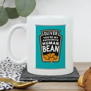 May include: White ceramic mug with a blue and gold design. The design features a can of beans with the text "Oliver You're My Favourite Human Bean".