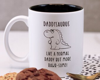 Può includere: Una tazza da caffè in ceramica bianca con un bordo nero. La tazza presenta un'illustrazione in bianco e nero di un dinosauro con il testo "Daddysaurus Like a normal daddy but more rawr-some!"