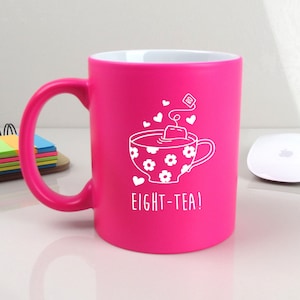 May include: A bright pink ceramic mug with a white design of a teacup with a teabag and the text "EIGHT-TEA!"
