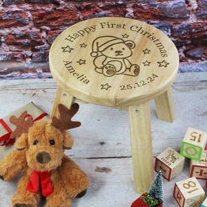 Peut inclure: Un petit tabouret en bois clair avec une assise ronde. L'assise est gravée de "Happy First Christmas", d'un ours en peluche portant un bonnet de Père Noël, d'étoiles, du nom "Amelia" et de la date "25.12.24". Un jouet renne en peluche et des blocs de jouets sont en arrière-plan.